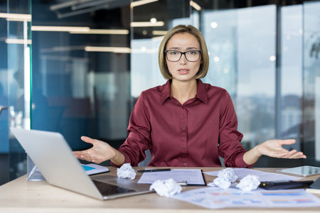 Businesswoman at her desk surrounded by crumpled papers and laptop, gesturing in frustration and confusion as she struggles with a stressful deadline and workplace pressureの写真素材