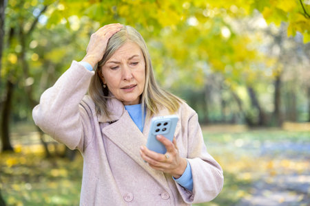 Senior woman in autumn park looks distressed and confused while reading her smartphone, holding her...