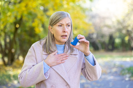 Female senior with an anxious expression experiencing an asthma attack in a park, holding an inhaler and placing a hand on her chest, seeking relief from breathing problemsの写真素材