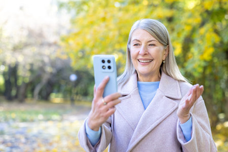 Senior woman is enjoying a video call on her smartphone in an autumn park, actively engaging and...