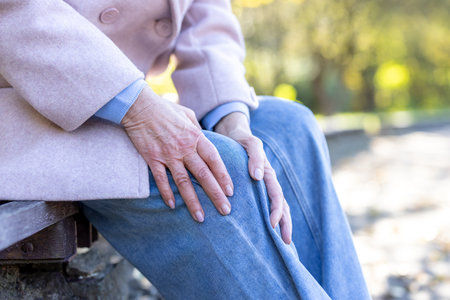 Senior woman feeling knee pain due to arthritis, gripping her leg while resting on a park bench, conveying discomfort and health challenges associated with agingの写真素材