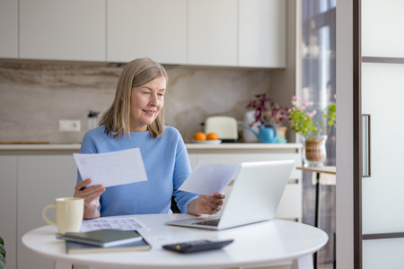 Senior woman at home managing finances, reviewing bills and documents at a desk with laptop and calculator, budgeting, planning retirement and household expenses with calm focusの写真素材