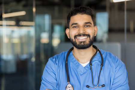Young man doctor smiling at the camera, confidently standing with a stethoscope around his neck, reflecting professionalism and dedicated healthcare service in a modern medical environmentの写真素材
