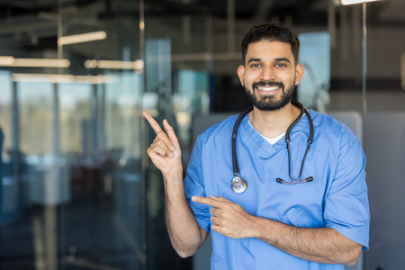 Young indian male doctor in blue scrubs and stethoscope smiling while using both hands pointing to the left, indicating good medical service and information in a modern hospital settingの写真素材