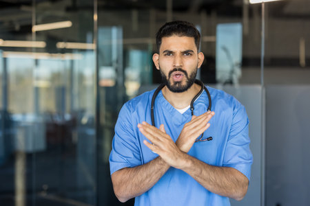 Young male doctor in blue scrubs with stethoscope crossing his arms in a clear stop gesture, serious expression signaling refusal, warning or objection in clinical settingの写真素材