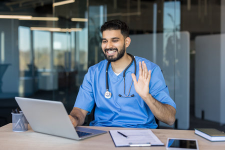 Male professional doctor wearing blue scrubs and stethoscope smiling, waving hand during virtual teleconsultation from an office, providing remote healthcare services to patientsの写真素材