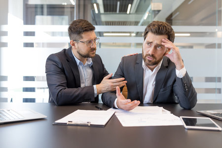 Businessman offering support and empathy to his sad, stressed colleague experiencing failure or a difficult financial problem, sitting together at a modern office desk discussing documentsの写真素材