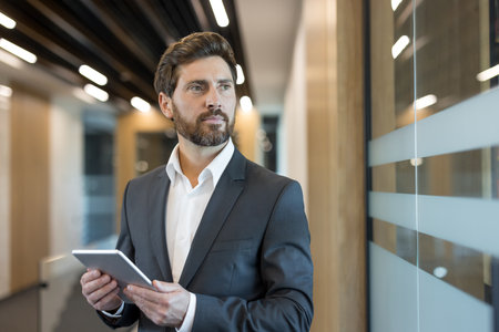 Businessman holding a digital tablet in a professional office environment, looking with focus and determination, representing corporate leadership and successの写真素材