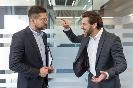 Two businessmen having an intense argument in a modern office, one executive shouting and pointing his finger at the other employee who is calmly listeningの写真素材