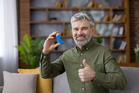 Senior grey-haired man sitting on a sofa in a modern living room, smiling and holding an asthma...