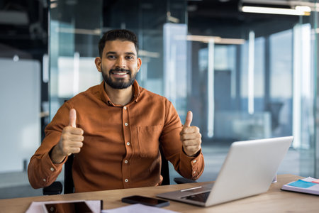 Indian businessman sitting at a modern office desk with a laptop, smiling confidently and giving a dual thumbs up, symbolizing approval, positivity, and achievement in his professional careerの写真素材