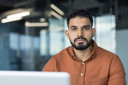 Indian businessman standing in a modern office, looking at camera with serious, confident expression smart-casual entrepreneur conveying professionalism, focus and leadershipの写真素材