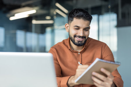Young businessman smiling, making notes in a notebook while working on a laptop in a modern office, focusing on his tasks and professional developmentの写真素材