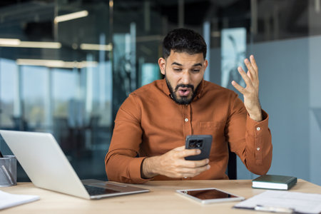 Businessman sitting at a desk in a modern office, reacting with surprise and shock while looking at his smartphone, receiving unexpected or disappointing newsの写真素材