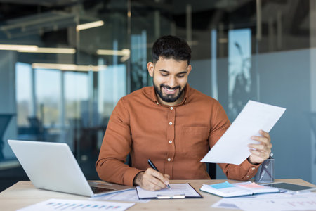 Young smiling businessman at his modern office desk signing documents, surrounded by laptop, charts and paperwork as he reviews financial data and plans strategyの写真素材