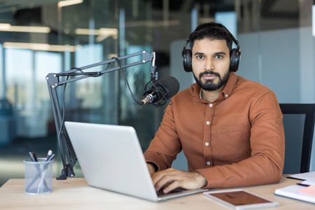 Young man with headphones and microphone typing on laptop in a modern office, recording and...