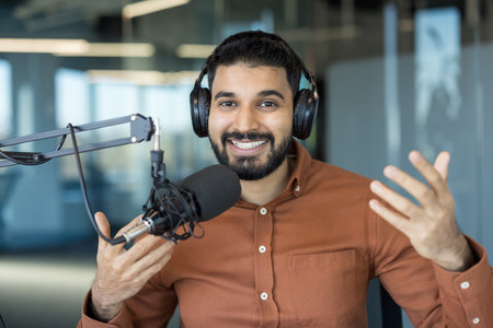 Young man wearing headphones and a rust-colored shirt, smiling and gesturing while talking into a professional microphone, actively creating audio content in a modern studio environmentの写真素材