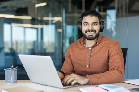 Young indian man in a modern office wearing headphones, smiling at camera while typing on a laptop,...
