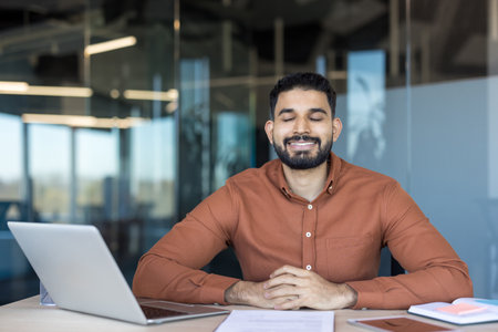 Young indian businessman seated at his desk with eyes closed and serene smile, practicing mindfulness and stress relief for calm, focus and well-being during a busy workdayの写真素材