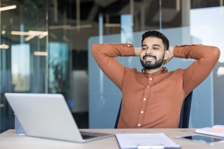 Young indian businessman sitting at office desk, hands behind head, smiling and looking satisfied after successfully completing work or taking a breakの写真素材
