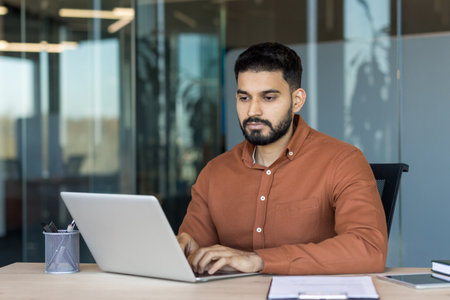 Young indian man staying focused while typing on a laptop at his desk, demonstrating dedication and productivity in a contemporary corporate environmentの写真素材