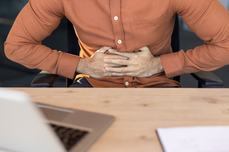Young professional clutching his abdomen in pain at an office desk, struggling with indigestion or stomachache while trying to work, stressed and feeling unwellの写真素材