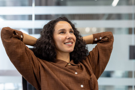 Young businesswoman with curly hair taking a comfortable break in the office, hands behind head, smiling contentedly and looking away, enjoying a moment of relaxationの写真素材