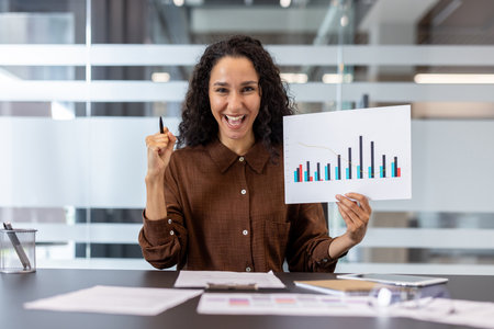 Mixed race businesswoman celebrating business success and achievement, holding a chart with increasing bars and a pen, expressing excitement while sitting at her office deskの写真素材