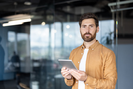 Young businessman holding a digital tablet with a serious expression, looking directly at the viewer, standing in a contemporary office representing digital communication and business technologyの写真素材