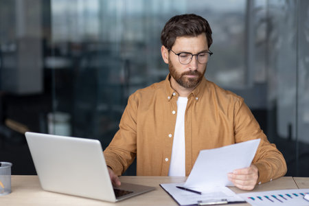 Businessman reading paper documents and reviewing reports while working on a laptop at a contemporary office desk, demonstrating dedication and success in his careerの写真素材
