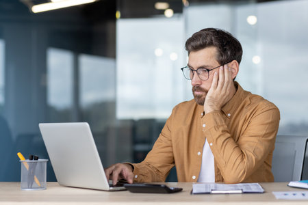 Businessman exhausted and bored at a desk, leaning on hand while typing on a laptop in a modern home office, showing stress, burnout and deadline pressureの写真素材