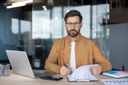 Businessman looking at camera, sitting at office desk, holding documents, writing on clipboard, with laptop and calculator in a modern office environmentの写真素材