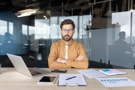 Serious male professional with a beard and glasses sitting at an office desk with a laptop, tablet, documents, and calculator, looking directly at the camera with arms crossedの写真素材