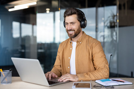 Happy bearded man in smart-casual attire wearing headphones, smiling at laptop during a virtual...