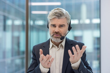 Mature bearded man communicating while wearing a headset and suit, providing call center customer service or online support in a modern office environmentの写真素材