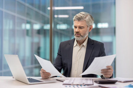 Mature businessman studying important paperwork, comparing financial reports and data analysis, making focused decisions while working in a modern corporate officeの写真素材