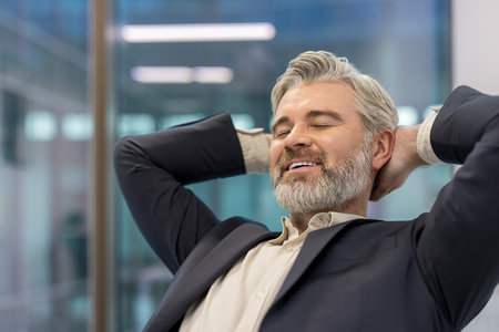 Mature businessman with gray hair and beard leaning back, eyes closed and smiling, feeling content and finding a moment of relaxation and satisfaction during a break at the officeの写真素材