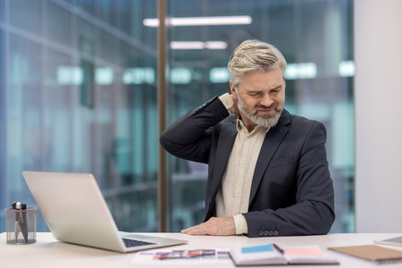 Mature businessman experiencing neck pain and discomfort while working on a laptop at his office desk, symbolizing work-related stress, health issues, and burnout from prolonged computer useの写真素材