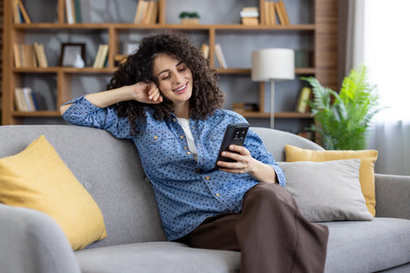 Woman with curly hair smiling and leaning back on a living room couch, enjoying leisure time while texting and browsing social media on a mobile phoneの写真素材