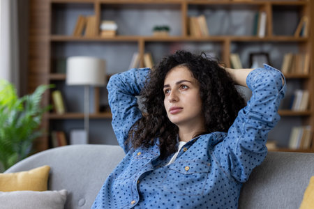 Young woman with curly hair reclining on a gray couch in a modern living room, hands behind head, looking away thoughtfully, relaxed and contemplative during a peaceful breakの写真素材