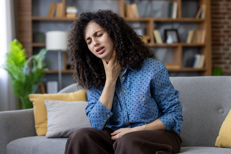 Young woman on a couch clutching her throat in pain, looking ill and fatigued at home, suffering...