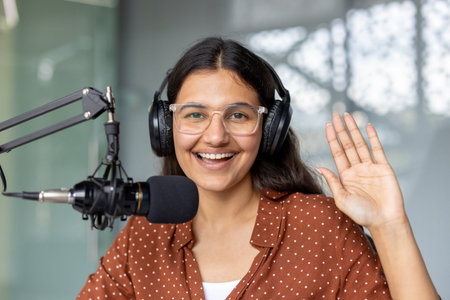 Indian woman wearing headphones and clear glasses, smiling and waving, recording a podcast in a modern studio with a professional microphone, creating engaging audio contentの写真素材