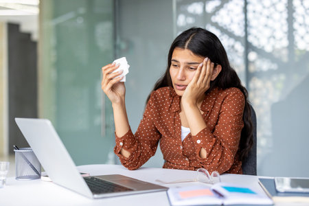 Stressed young indian woman at her laptop in a modern office, holding a tissue and crying while overwhelmed by work pressure, burnout and emotional breakdownの写真素材