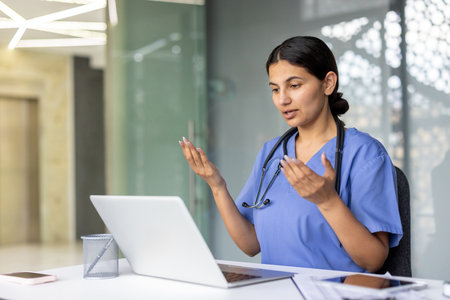 Young female medical professional wearing scrubs and stethoscope providing online healthcare services and remote medical advice during a virtual consultation on a laptopの写真素材