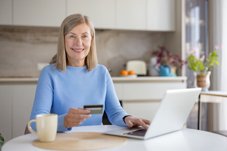 Smiling senior woman in a cozy kitchen using a laptop and holding a credit card for online shopping and banking, relaxed at the counter with a mug and natural lightの写真素材