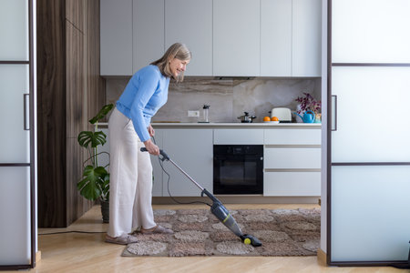 Senior woman vacuuming carpet in a modern kitchen, smiling as she maintains a clean home, engaged in everyday domestic chores and enjoying active, comfortable retirement routineの写真素材