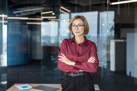 Confident businesswoman with a bob haircut and glasses, wearing a maroon shirt, stands with crossed arms, looking determined inside a contemporary office workplaceの写真素材