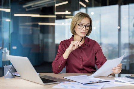 Businesswoman reviewing financial reports and data on paper, concentrating on her work at a desk with a laptop and various documents in a contemporary office environmentの写真素材