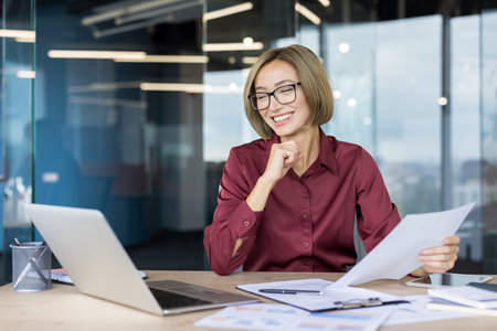 Young businesswoman smiling, holding documents and working on a laptop at a bright office desk, celebrating work success, development, and a positive career journeyの写真素材