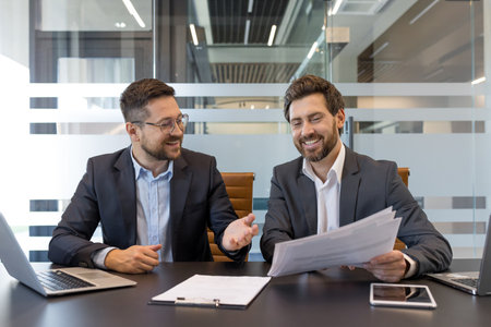 Two smiling businessmen sitting at a modern office table, discussing work documents and collaborating happily during a corporate meeting with laptops and a tablet on the deskの写真素材